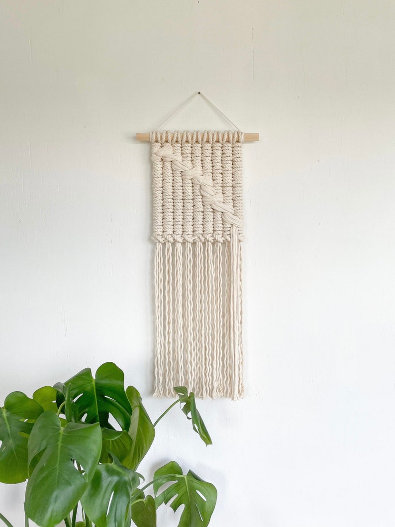 May include: A white macrame wall hanging with a woven pattern and fringe. The hanging is attached to a wooden dowel rod.