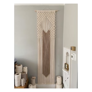 May include: A large macrame wall hanging with a geometric design in cream and brown. The hanging is made with natural fibers and has a fringe at the bottom.