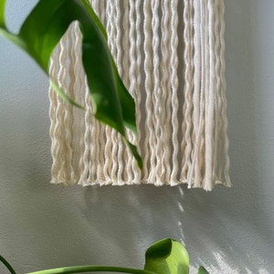 May include: A white macrame wall hanging with a fringe bottom. The hanging is made of thick, twisted cotton cord.