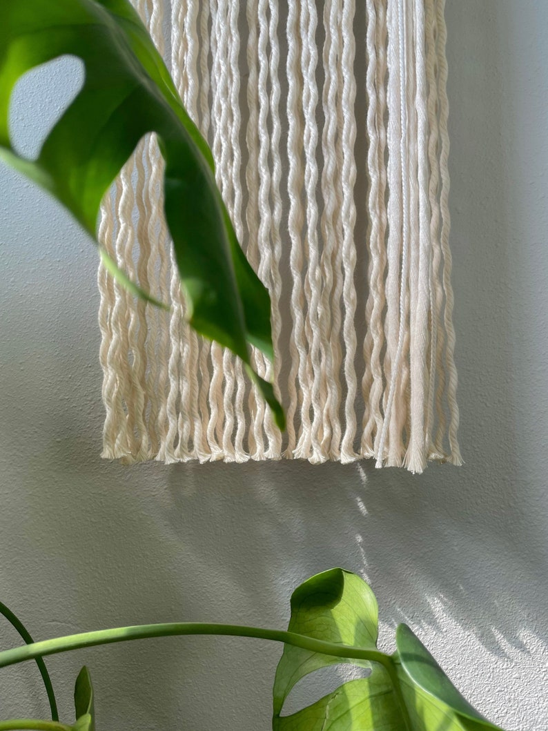 May include: A white macrame wall hanging with a fringe bottom. The hanging is made of thick, twisted cotton cord.