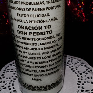 Puede incluir: Una vela de oraci&oacute;n blanca con texto negro, con las palabras "DON PEDRITO" y una oraci&oacute;n en espa&ntilde;ol e ingl&eacute;s. La vela est&aacute; sobre un mantelito de encaje blanco, con un fondo oscuro.