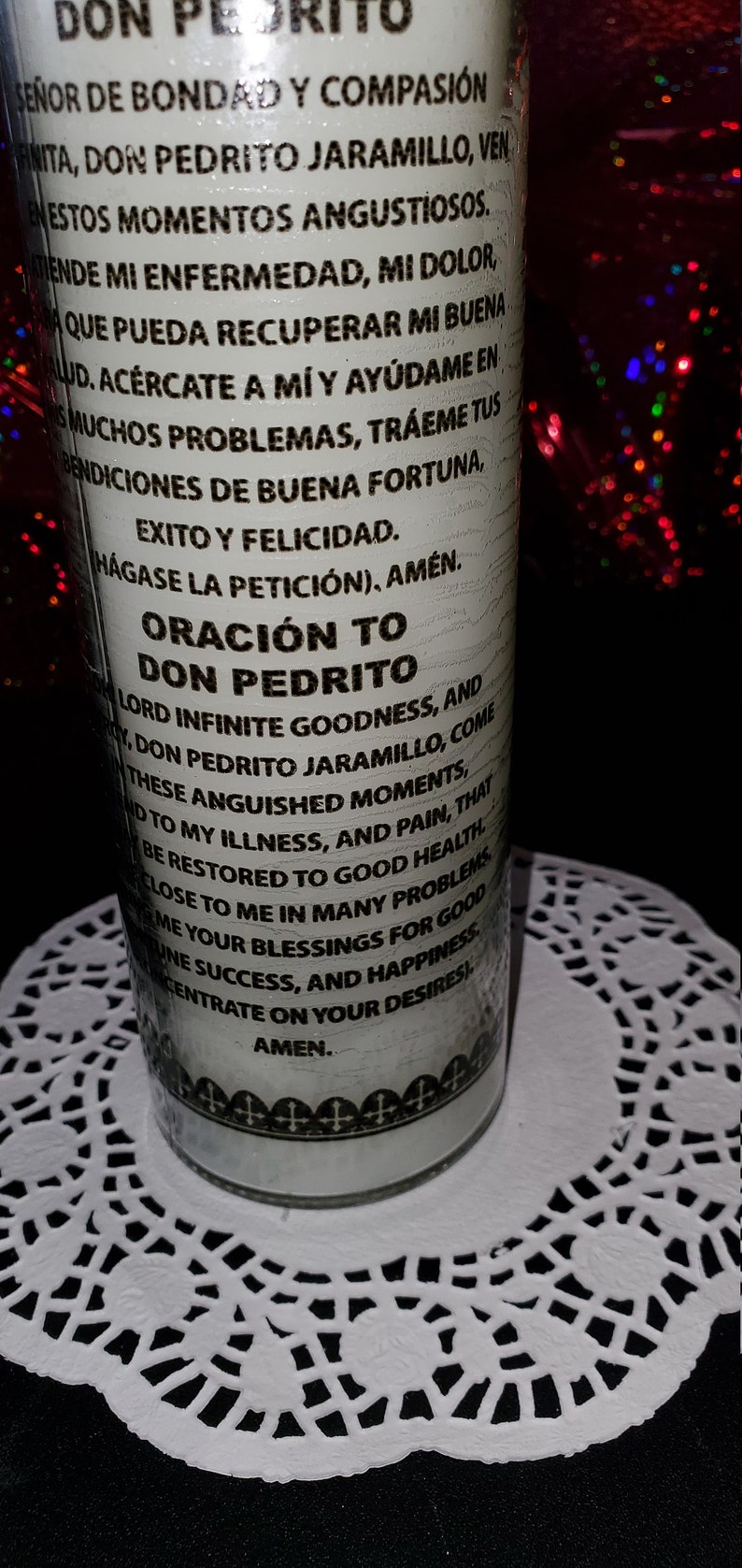 Puede incluir: Una vela de oraci&oacute;n blanca con texto negro, con las palabras "DON PEDRITO" y una oraci&oacute;n en espa&ntilde;ol e ingl&eacute;s. La vela est&aacute; sobre un mantelito de encaje blanco, con un fondo oscuro.