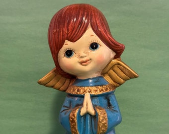 May include: A ceramic angel figurine with red hair, blue wings, and a blue dress with gold trim. The angel is praying with its hands clasped together.