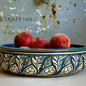 May include: A ceramic bowl with a blue rim and a white and blue leaf pattern holds several ripe peaches. The peaches are a vibrant red color with a fuzzy texture. The bowl is sitting on a white surface.