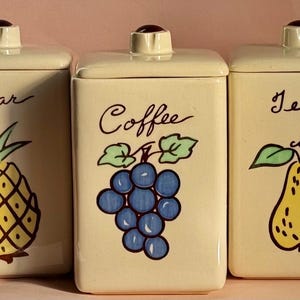 May include: Three vintage ceramic canisters with lids, each featuring a different fruit design. The canister on the left has a pineapple and says "Sugar". The middle canister has grapes and says "Coffee". The canister on the right has a pear and says "Tea".