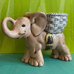 May include: A ceramic elephant planter with a brown and white wicker basket on its back. The elephant is light brown with a yellow saddle and a brown strap.