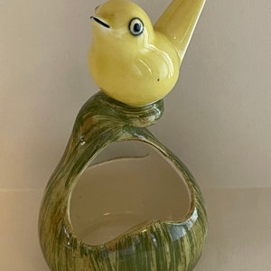 May include: A yellow ceramic bird perched on a green and white ceramic planter with a curved opening. The planter has a woodgrain pattern.