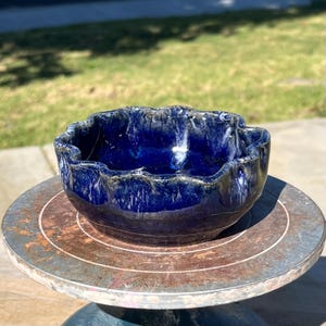May include: A handmade ceramic bowl with a wavy rim, glazed in a deep blue color with white streaks. The bowl sits on a round, rustic, metal surface. The bowl's interior is a darker shade of blue.