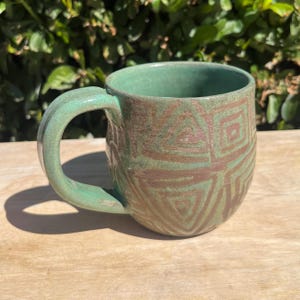May include: A handmade ceramic mug with a green glaze and brown geometric patterns. The mug has a rounded shape with a large handle. The mug is sitting on a wooden surface.