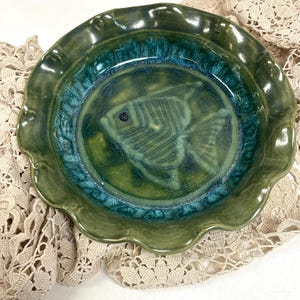 May include: A handcrafted ceramic plate with a fish design. The plate features a scalloped edge and is glazed in shades of green and teal. The fish is etched into the center of the plate.