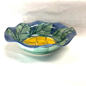 May include: A hand-painted ceramic bowl with a wavy rim. The bowl features a design of green leaves and blue accents around the rim, with a yellow interior. The bowl has a light green base.