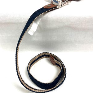 Puede incluir: Un cintur&oacute;n tejido en azul marino y beige con hebilla plateada y detalles en cuero marr&oacute;n. El cintur&oacute;n presenta un dise&ntilde;o a rayas. El cintur&oacute;n es nuevo y tiene una etiqueta adjunta. El cintur&oacute;n mide unos 120 cm de largo.