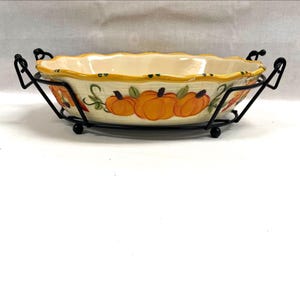 May include: An oval ceramic baking dish with a scalloped rim, featuring a painted pattern of orange pumpkins and green vines on a cream background, held within a black metal wire frame stand with four small ball feet for stability.