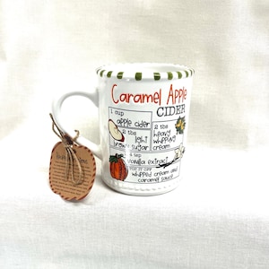 May include: A white ceramic mug with a green striped rim featuring a printed recipe for Caramel Apple Cider. The mug includes illustrations of an apple slice and a pumpkin, with a brown apple-shaped tag tied to the handle with twine.