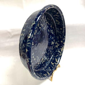 May include: A deep blue ceramic bowl with a speckled pattern of cream-colored spots. The bowl has a rounded shape with a slightly raised rim. The bowl is displayed on a gold-colored stand.