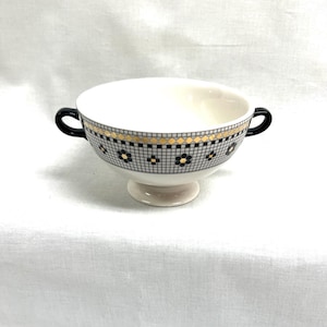 Pu&ograve; includere: Ciotola per zuppa in ceramica bianca con manici neri. La ciotola presenta una fascia decorativa con un motivo geometrico nero e oro e accenti floreali neri. Poggia su una piccola base rotonda.
