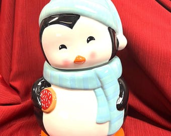 Puede incluir: Un tarro de galletas de cerámica con forma de pingüino que lleva un gorro de punto azul claro y una bufanda a rayas a juego. El pingüino tiene la barriga blanca, las alas negras y los pies naranjas, y sostiene una galleta con chispas rojas.