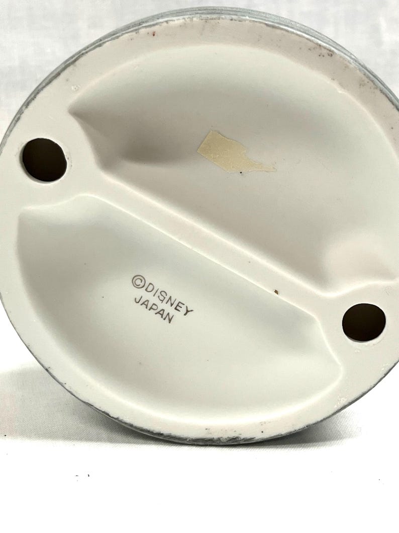 Puede incluir: Parte inferior de una figura de cer&aacute;mica blanca con dos agujeros y una ranura central. La base tiene un borde gris y el texto "&copy;DISNEY JAPAN". Tambi&eacute;n hay una peque&ntilde;a pegatina descolorida.