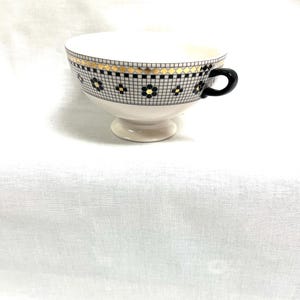 Pu&ograve; includere: Una tazza da t&egrave; bianca con un manico nero e una fascia decorativa con un motivo geometrico nero e oro. La tazza da t&egrave; poggia su una piccola base.