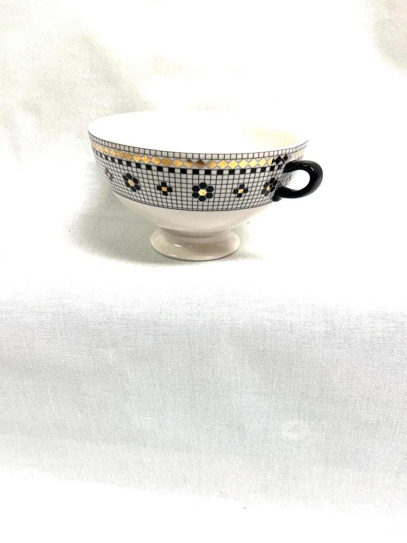 Pu&ograve; includere: Una tazza da t&egrave; bianca con un manico nero e una fascia decorativa con un motivo geometrico nero e oro. La tazza da t&egrave; poggia su una piccola base.