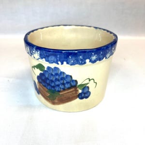 May include: A ceramic bowl with a cream-colored base and a thick, hand-painted blue rim. The exterior features a painted design of a cluster of blue grapes resting in a brown basket with green leaves, rendered in a simple, folk-art style.