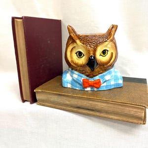 May include: A ceramic owl figurine featuring brown feathers, yellow eyes, and a black beak. The owl wears a light blue and white checkered shirt with a bright orange bow tie. The figurine is positioned on top of a brown book next to a maroon book.