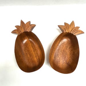 May include: Two wooden pineapple-shaped bowls. The bowls are a warm brown color with a natural wood grain. The top of each bowl features a detailed, carved pineapple crown. These decorative bowls are ideal for serving snacks or as tropical decor.
