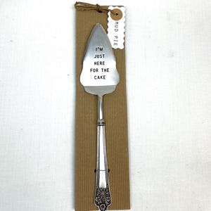 May include: A silver cake server with the text "I'M JUST HERE FOR THE CAKE" stamped on the blade. The server is attached to a brown cardboard backing with a "MUD PIE" tag, tied with twine. The handle has an ornate design.