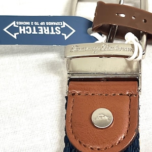 Puede incluir: Primer plano de un cintur&oacute;n Tommy Bahama con hebilla plateada y detalles en cuero marr&oacute;n. El cintur&oacute;n tiene una tela tejida azul marino y una etiqueta que dice "STRETCH SE EXTIENDE HASTA 5 cm."