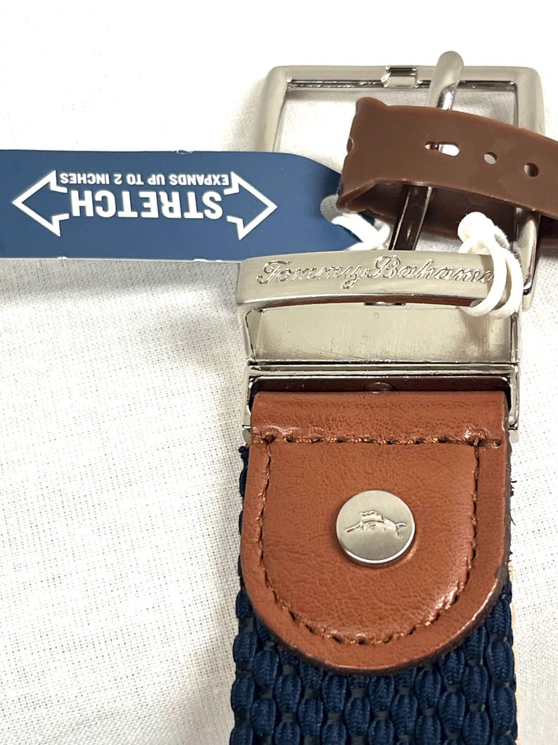 Puede incluir: Primer plano de un cintur&oacute;n Tommy Bahama con hebilla plateada y detalles en cuero marr&oacute;n. El cintur&oacute;n tiene una tela tejida azul marino y una etiqueta que dice "STRETCH SE EXTIENDE HASTA 5 cm."