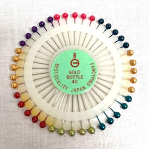 May include: A circular set of sewing pins with a white plastic holder. The pins have colorful ball heads in shades of red, purple, gold, green, and teal. The center of the holder has a green label with the text "GOLD BOTTLE 40 FANCY PINS JAPAN".