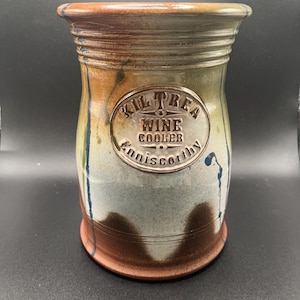 May include: A handcrafted ceramic wine cooler with a cylindrical shape. The cooler features a textured surface with a blend of brown, orange, and blue hues. The text "Kiltrea Wine Cooler Enniscorthy" is embossed on the front.