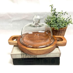 May include: A wooden cheese board with a clear glass dome. The board has a round base with two handles and a raised edge. The dome has a small handle on top. The board is sitting on top of two books.