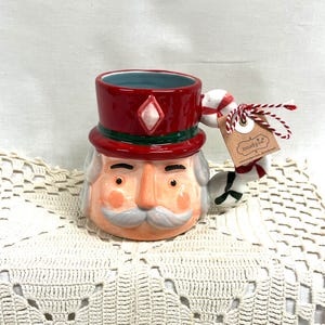 May include: A ceramic nutcracker mug featuring a red hat with a diamond accent, a painted face with a gray mustache, and a handle shaped like a candy cane. A brown paper tag with the text mudpie is attached to the handle with red and white string.