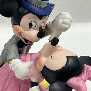 Puede incluir: Una figura de Mickey Mouse y Minnie Mouse bailando. Mickey lleva un traje gris, un sombrero de copa azul y guantes blancos. Minnie lleva un vestido y un lazo rosas. La figura tiene un estilo cl&aacute;sico de Disney.