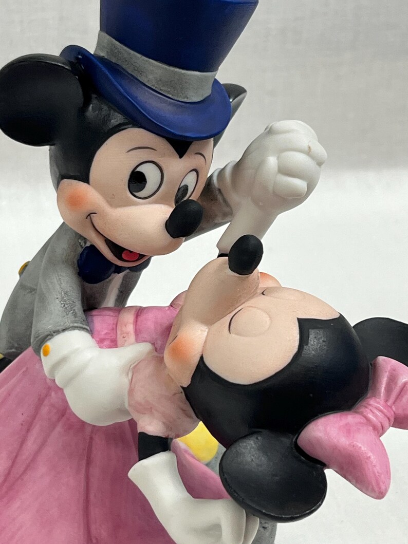 Puede incluir: Una figura de Mickey Mouse y Minnie Mouse bailando. Mickey lleva un traje gris, un sombrero de copa azul y guantes blancos. Minnie lleva un vestido y un lazo rosas. La figura tiene un estilo cl&aacute;sico de Disney.