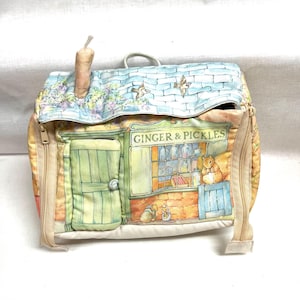 May include: A fabric backpack featuring a printed illustration of a shop storefront labeled GINGER & PICKLES. The design includes a light green door, a shop window with jars, and small animal characters. The top flap has a blue brick pattern.