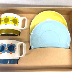 May include: A set of two white ceramic mugs with floral designs in yellow, blue, and orange, and two matching plates in yellow and light blue. The mugs have a stacked design and are presented in a cardboard box.