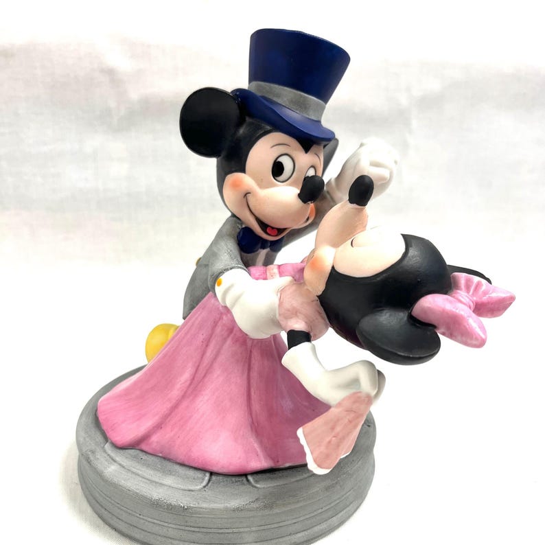 Puede incluir: Figura de cer&aacute;mica de Mickey Mouse y Minnie Mouse bailando. Mickey lleva un traje gris, un sombrero de copa azul y una pajarita. Minnie lleva un vestido y un lazo rosas. La base es una plataforma circular gris.