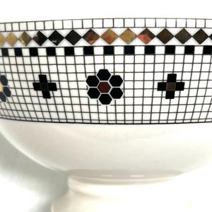 Pu&ograve; includere: Una ciotola in ceramica bianca con un motivo a mosaico decorativo. Il mosaico presenta forme a diamante nere, dorate e marroni, croci nere e un motivo floreale. La ciotola ha una base a piedistallo.