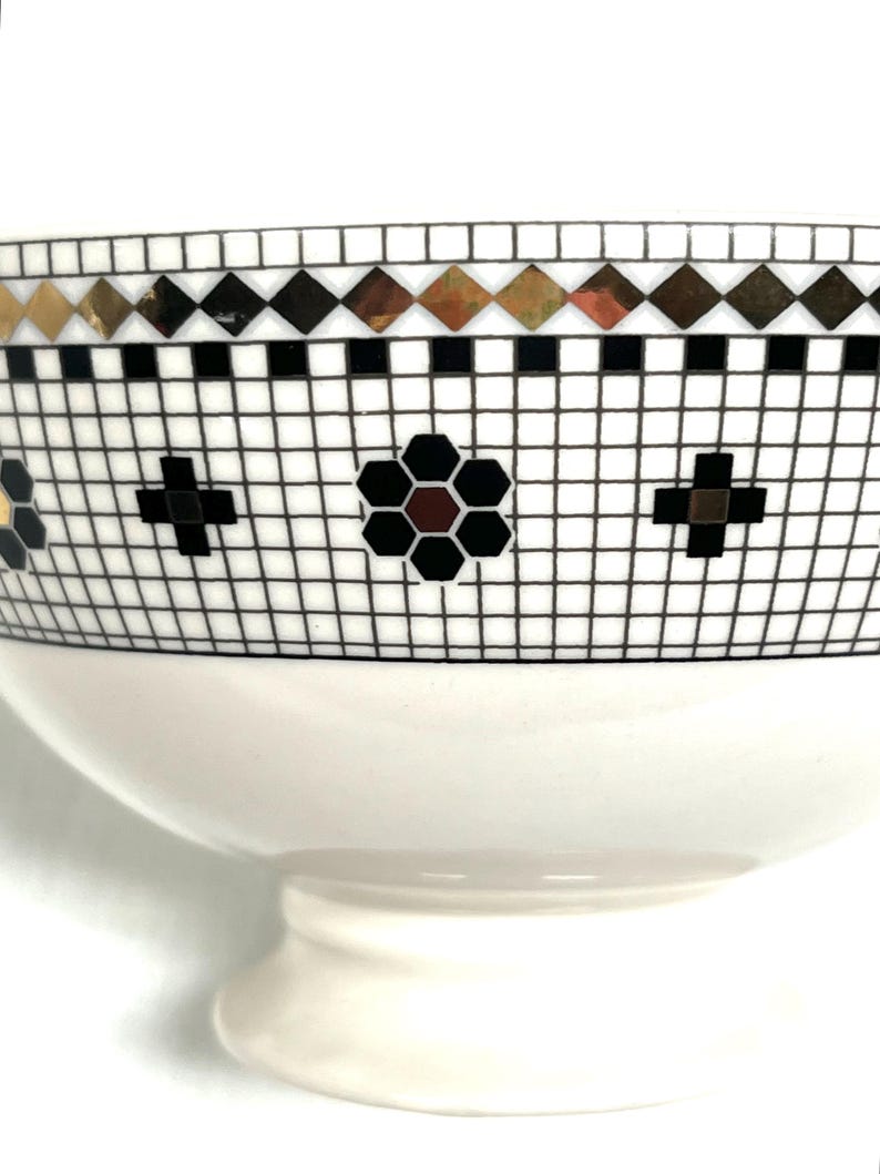Pu&ograve; includere: Una ciotola in ceramica bianca con un motivo a mosaico decorativo. Il mosaico presenta forme a diamante nere, dorate e marroni, croci nere e un motivo floreale. La ciotola ha una base a piedistallo.