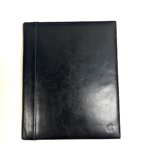 May include: A rectangular black portfolio folder with a smooth, dark finish. The item features a vertical stitched seam along the left side and a small, embossed Apple logo located in the bottom right corner of the front cover surface.