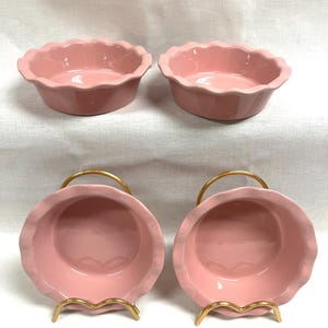 May include: Four pink ceramic bowls with scalloped edges. Two bowls are displayed upright, and two are in gold-colored metal stands. The bowls have a smooth, glossy finish. The background is a white fabric.
