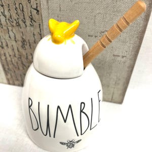 May include: A white ceramic honey pot with a lid featuring a yellow bee topper. The word BUMBLE is printed in black lettering on the front, accompanied by a small black bee illustration. A light brown wooden honey dipper rests inside the pot.