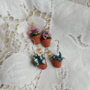 May include: Handmade earrings featuring miniature terracotta pots with sculpted flowers and foliage. The earrings have gold-colored hooks and decorative beads. The flowers are pink and the foliage is green and white.