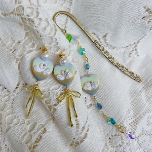 May include: A collection of swan-themed jewelry and accessories. Includes a pair of heart-shaped earrings with gold bows, a matching pendant, and a decorative bookmark. The earrings and pendant feature a swan design with pearl accents.
