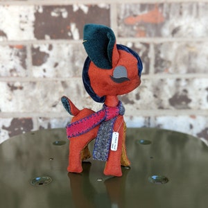 Può includere: Un cervo di feltro rosso e marrone con un cappello blu e verde e una sciarpa rossa e blu. Il cervo indossa un'etichetta che dice "Made This".