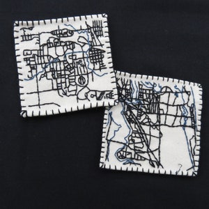 May include: Two white fabric squares with black stitched outlines of maps. The maps are of different areas, but both have a blue river or stream running through them.