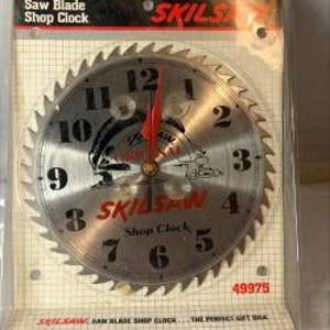 May include: A circular Skilsaw shop clock featuring a metallic saw blade face with black numbers 1 through 12, red clock hands, and the Skilsaw logo. The clock is encased in clear plastic packaging with a grid-patterned backing card and text.