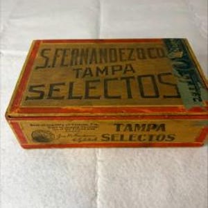 May include: A rectangular wooden cigar box with a natural wood grain top featuring the black printed text S. FERNANDEZ & CO. TAMPA SELECTOS. The sides are painted in red and yellow with additional black text and a circular logo on the front panel.